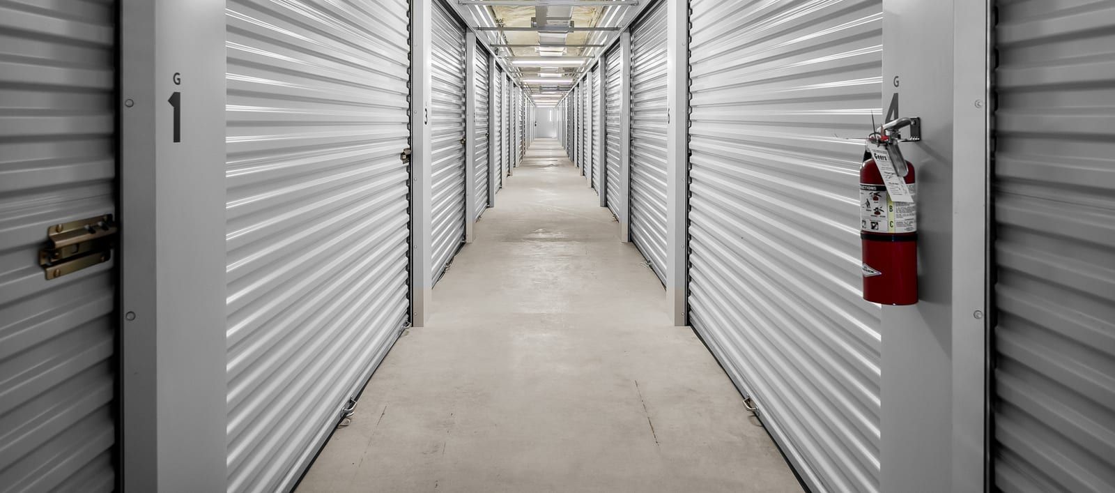 Indoor storage hallway lined with gray roll-up unit doors, with lights and ductwork along the ceiling