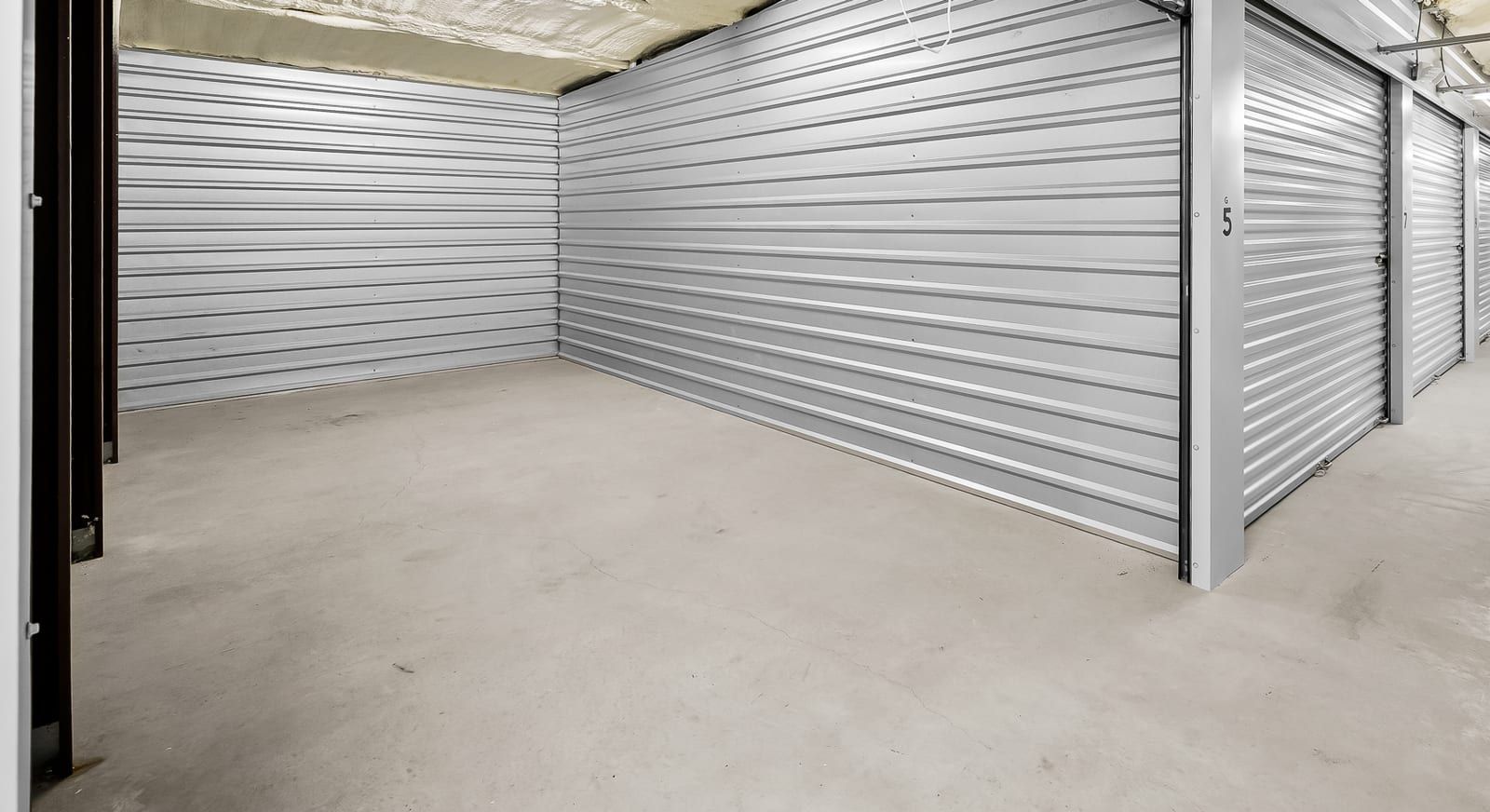 Open, empty storage unit with steel walls and a concrete floor beside a long indoor hallway of roll-up doors