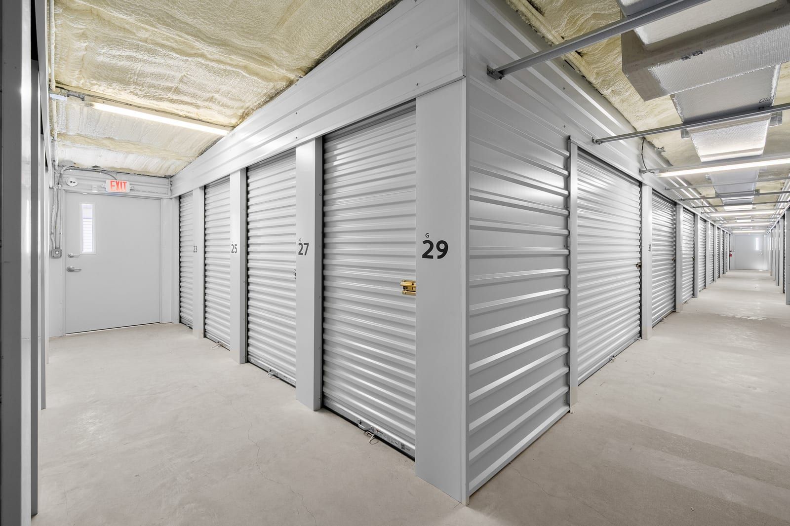 Bright indoor hallway corner lined with numbered white roll-up storage doors and an exit door