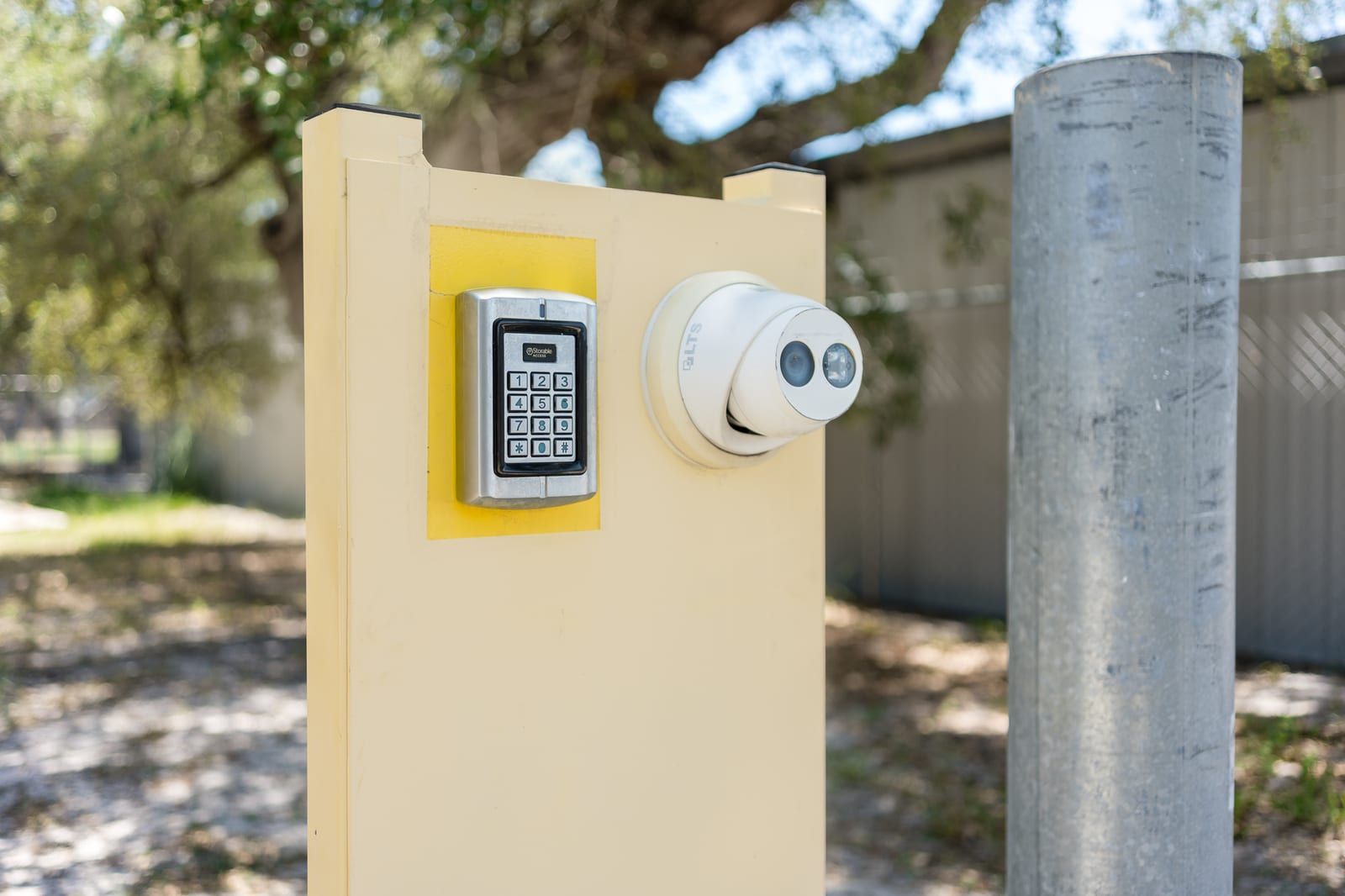 Close-up of a gate entry keypad and a white dome security camera mounted on a yellow post