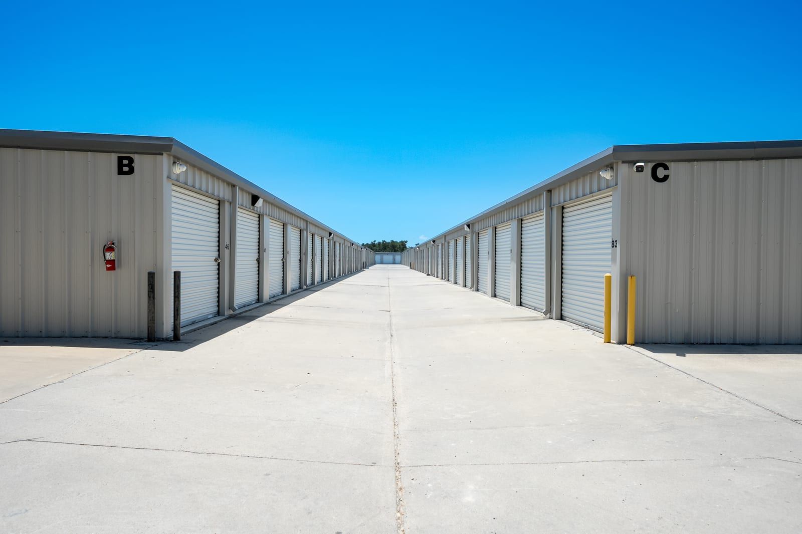 Wide concrete drive aisle between storage buildings B and C, lined with white roll-up doors under a clear blue sky