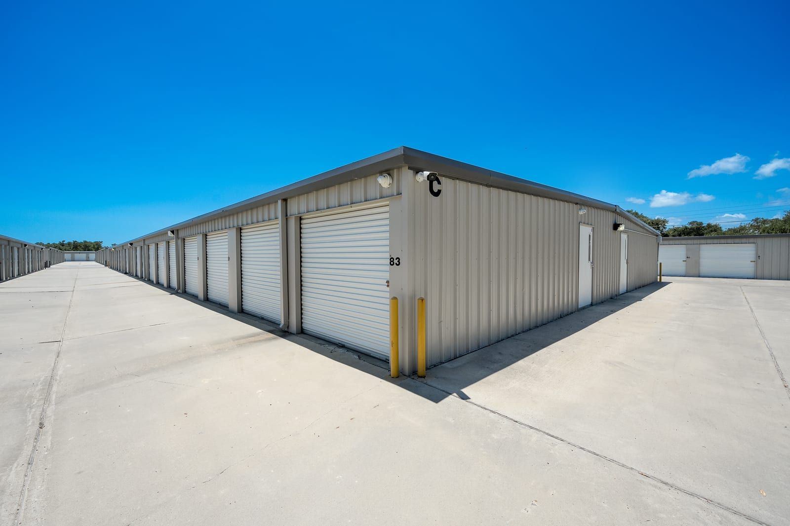 Corner of a tan metal storage building with white roll-up doors, two wall-mounted cameras and a wide concrete drive