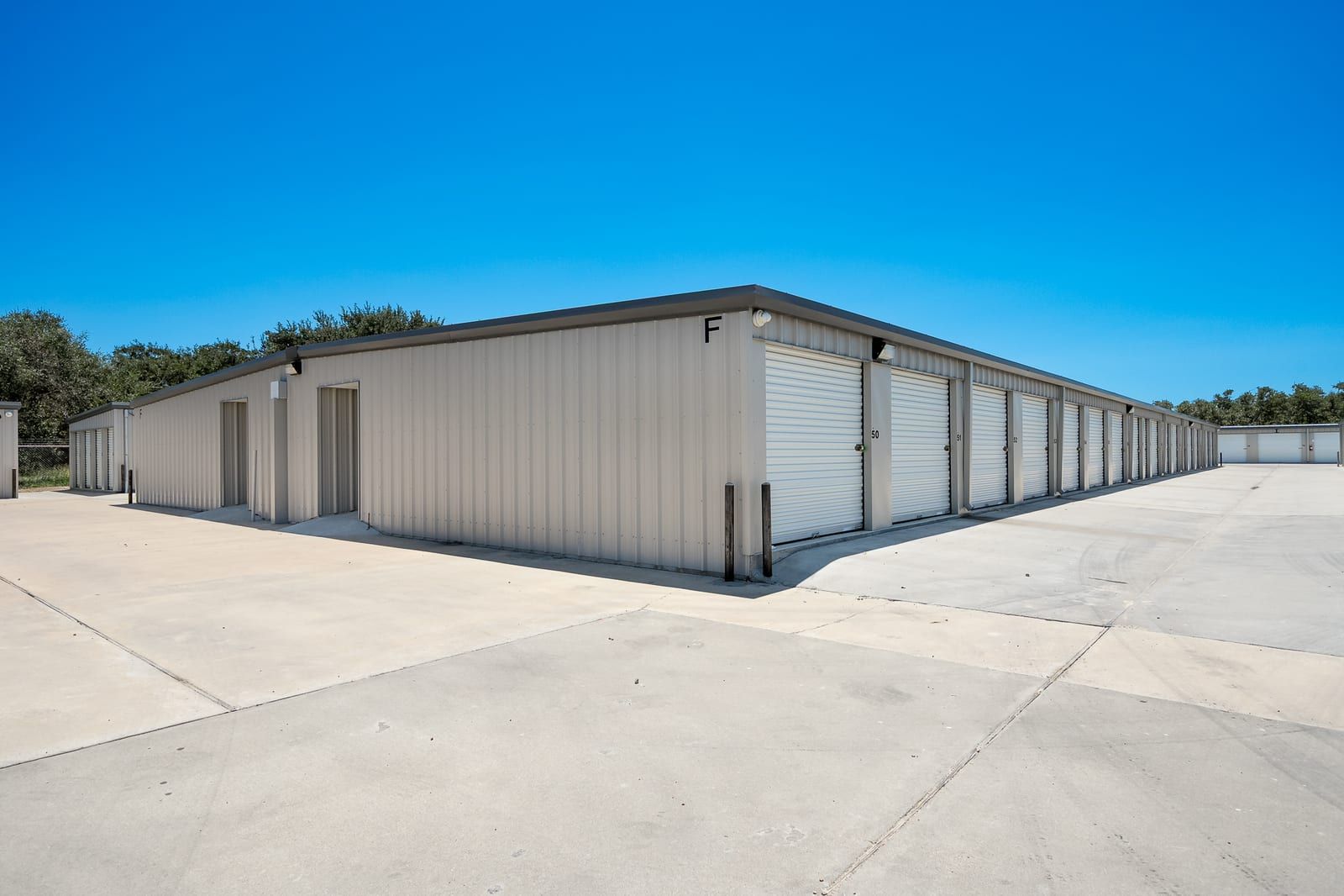 Long row of white roll-up storage doors on a tan metal building beside a wide concrete drive, trees behind